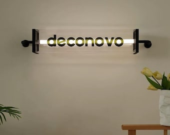 May include: A modern wall-mounted light fixture with the word "deconovo" in black lettering. The light is encased in a clear rectangular tube, supported by black metal brackets on each end. The fixture emits a warm, inviting glow.