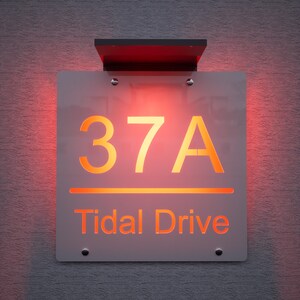 以下が含まれることがあります： オレンジ色の光で「37A」の数字と「Tidal Drive」の文字が表示された、照明付きの住所看板。看板は質感のある灰色の壁に取り付けられており、黒い上部が付いています。
