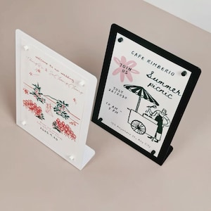 May include: Two acrylic sign holders, one white and one black, each displaying a printed card. The white holder features wedding text and floral illustrations. The black holder displays a Cafe Rimberio summer picnic event flyer with a food cart graphic.