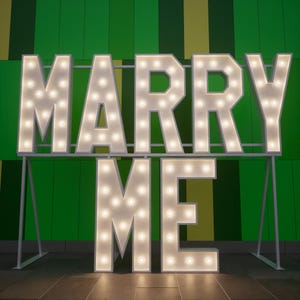 May include: Large white marquee letters spelling "MARRY ME" with illuminated bulbs. The letters are mounted on a white metal frame against a green and yellow background. The letters are lit up, creating a bright and festive display.