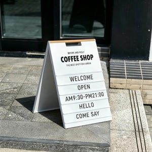Pode incluir: Placa em forma de A branca para uma cafeteria, com texto preto. A placa diz "COFFEE SHOP" e "THE BEST SPOT FOR A BREW". O texto adicional inclui "WELCOME", "OPEN", "AM9:30-PM21:00", "HELLO" e "COME SAY".