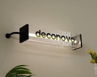 May include: A rectangular, clear acrylic light fixture with the word "deconovo" in black lettering. The fixture has a black metal frame and a curved arm attached to a wall. A white light is visible inside the fixture.