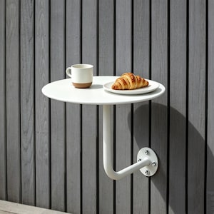 Puede incluir: Una mesa redonda blanca montada en la pared con un brazo de soporte curvo. Una taza blanca con base marrón y un plato con un cruasán están sobre la mesa. La mesa está montada en una pared de madera gris.