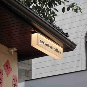 May include: A white rectangular sign with black text that reads "peekaboo coffee" is mounted on a brown awning. The sign is illuminated from within.