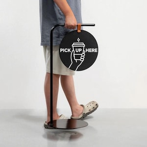 Puede incluir: Un letrero de metal negro con un diseño circular que dice "PICK UP HERE" en texto blanco. El letrero presenta un gráfico blanco de una mano sosteniendo una taza de café. El letrero está adjunto a un soporte de metal negro.