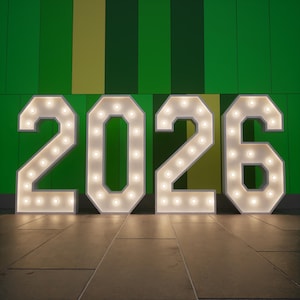 Puede incluir: Grandes letras de marquesina iluminadas que deletrean el año "2026". Cada letra blanca está delineada con pequeñas bombillas de tonos cálidos. Las letras se alzan sobre un suelo embaldosado contra un fondo de paneles verticales verdes y amarillos.