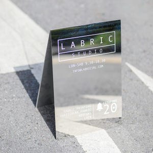 Op de afbeelding: Een spiegelbord met de tekst "LABRIC STUDIO" in het wit. Het bord bevat ook de openingstijden, contactgegevens en aangeboden diensten: stof bedrukken, bijzondere stof, knopen en kant. Een belpictogram met het nummer 20 is ook zichtbaar.