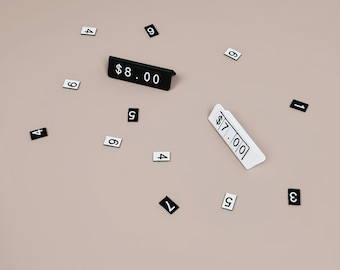 May include: Two angled price display blocks, one black with white text reading $8.00 and one white with black text reading $7.00, surrounded by scattered small rectangular tiles featuring individual black and white printed numbers.