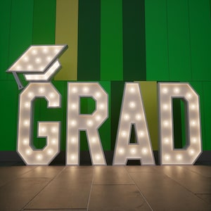 Peut inclure: Grandes lettres lumineuses épelant "GRAD" avec une coiffe de graduation. Les lettres et la coiffe sont blanches avec de nombreuses petites lumières aux tons chauds. Le fond est un mur rayé vert et jaune, et le sol est carrelé.