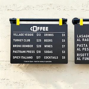 May include: A black rectangular menu board with white text reading COFFEE, VILLAGE VEGGIE, TURKEY CLUB, BRONX BOMBER, PASTRAMI PRESS, SPICY ITALIANO, DRINKS, BEERS, WINES, SODAS, COCKTAILS, and 11am to 3pm - Ask for today's specials, hung by yellow clips.