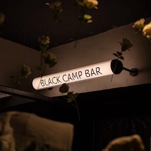 May include: A glowing, rectangular sign with the words "BLACK CAMP BAR" in white lettering. The sign is mounted on a wall and surrounded by hanging flowers. The overall aesthetic is dark and moody.