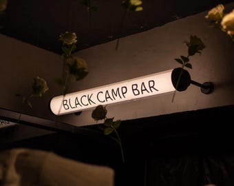 May include: A rectangular, illuminated sign with the words "BLACK CAMP BAR" in white lettering. The sign is mounted on a wall, and the background is dark. Artificial flowers hang from the ceiling.