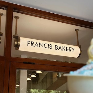 以下が含まれることがあります： 木製のドアの上に「FRANCIS BAKERY」の照明付き看板が吊り下げられています。看板は長方形で端が丸く、テキストは太字のサンセリフ体です。看板は真鍮色のロッドで吊り下げられています。