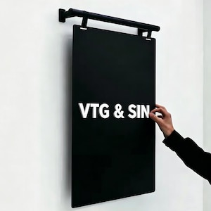 May include: A black rectangular sign with the white text "VTG & SIN" is mounted on a white wall. The sign is suspended from a black metal bar. A hand is touching the sign.