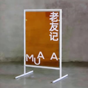 May include: A white metal sign with an orange translucent panel. The sign features white Chinese characters and the letters "MUA" in a modern font. The sign is free-standing with a rectangular frame and two supporting legs.