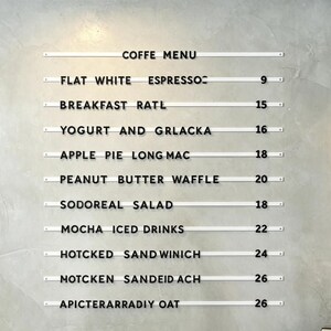 May include: A coffee menu displayed on a gray wall. The menu includes items like Flat White Espresso, Breakfast Ratl, Yogurt and Grlacka, Apple Pie Long Mac, Peanut Butter Waffle, Sodoreal Salad, Mocha Iced Drinks, and more.