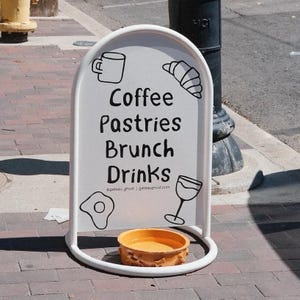 May include: A white, arched sandwich board sign with the words "Coffee, Pastries, Brunch, Drinks" in black. The sign has illustrations of a coffee mug, croissant, fried egg, and wine glass. A small, orange bowl sits at the base.