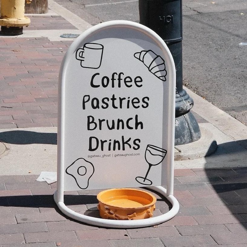 May include: A white, arched sandwich board sign with the words "Coffee, Pastries, Brunch, Drinks" in black. The sign has illustrations of a coffee mug, croissant, fried egg, and wine glass. A small, orange bowl sits at the base.