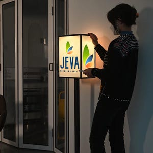 以下が含まれることがあります： 「JEVA PREMIUM QUALITY」の文字と、緑、青、オレンジの葉のデザインが施されたロゴが入った、正方形の照明付き看板。看板は黒いポールに取り付けられています。人が看板を持っています。