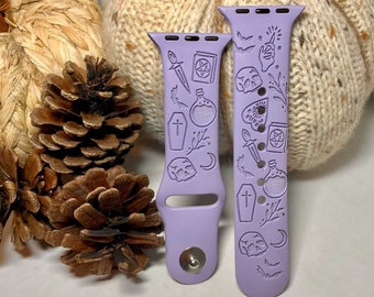 May include: Two lavender silicone watch bands with engraved Halloween designs. One band features a skull, crescent moon, coffin, potion bottle, and bats. The other band features a skull, crescent moon, coffin, potion bottle, dagger, pentagram, and a hand holding a wand.