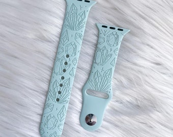 May include: Two light blue silicone Apple Watch bands with a crystal design. The bands are shown on a white fuzzy surface.