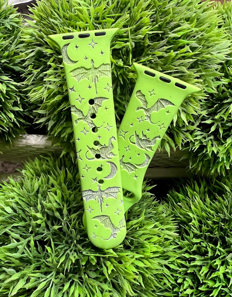 May include: A pair of green Apple Watch bands with a bat and moon design. The bands are made of silicone and have a smooth finish.