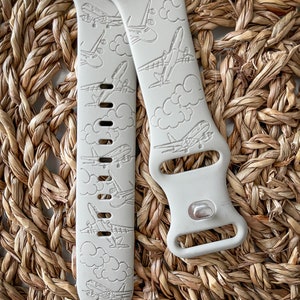 May include: A pair of light gray silicone watch bands with a repeating pattern of airplanes and clouds. The bands are designed for Apple Watch.