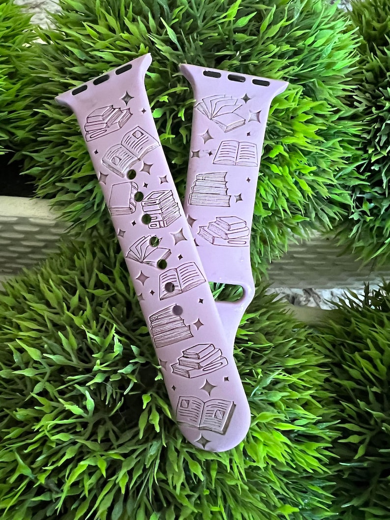 May include: A lavender-colored Apple Watch band with a white book and stack of books design. The band has a repeating pattern of books and stars.