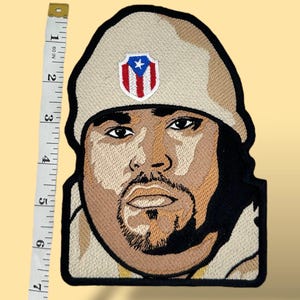 May include: Embroidered patch featuring a portrait of a man wearing a tan hat. The patch includes a shield with the Puerto Rican flag. The patch measures approximately 6 inches in height.