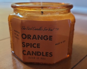 May include: A brown glass jar candle with a label that reads "The Best Candles For You! TM" and "ORANGE SPICE CANDLES" with the text "MADE IN USA" at the bottom. The label is a light brown color with black text.