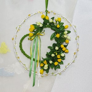 Könnte beinhalten: Ein kreisförmiger Blumenkranz mit gelben und weißen tulpenartigen Blüten und grünen Blättern. Der Kranz hat eine grüne Schleife und Bänder und wird von einem grünen Band gehalten. Der Kranz ist mit kleinen, klaren Perlen verziert.