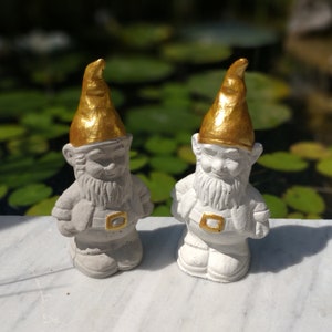 May include: Two white concrete garden gnomes with gold painted hats. The gnomes are standing on a white surface.