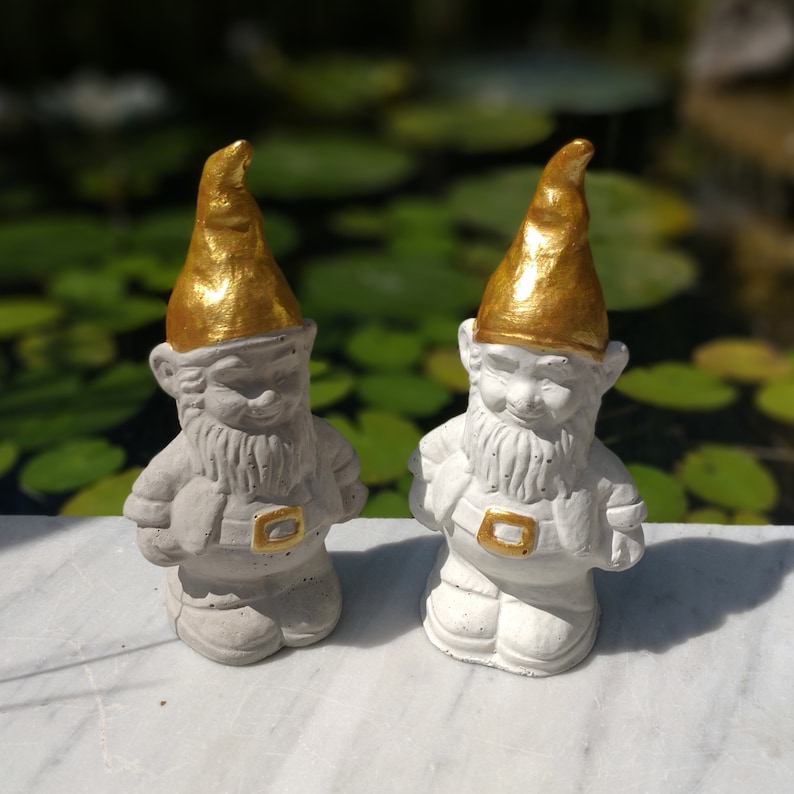 May include: Two white concrete garden gnomes with gold painted hats. The gnomes are standing on a white surface.