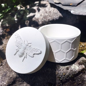 Könnte beinhalten: Weiße Keramikdose mit einem Wabenmuster und einem Deckel mit einem erhabenen Bienendesign.