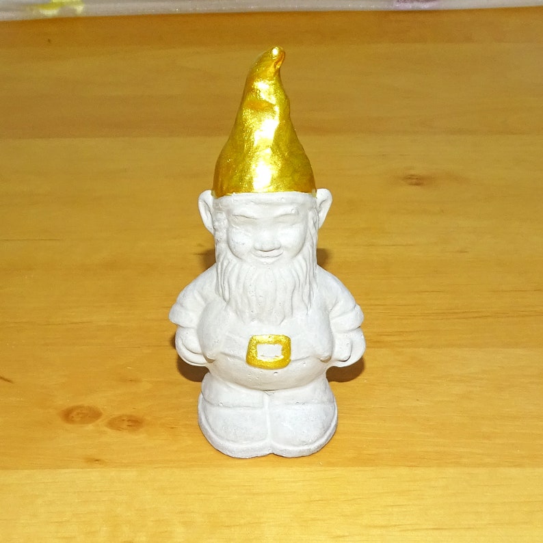 May include: A white garden gnome figurine with a gold painted hat. The gnome has a white beard and is wearing a brown belt.