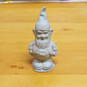 May include: A white ceramic garden gnome figurine with a pointed hat, a beard, and a belt. The gnome is smiling and has a friendly expression.