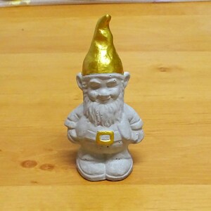 May include: A white ceramic garden gnome with a gold painted hat. The gnome has a white beard and a friendly smile.