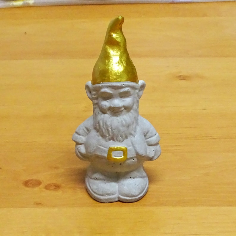 May include: A white ceramic garden gnome with a gold painted hat. The gnome has a white beard and a friendly smile.