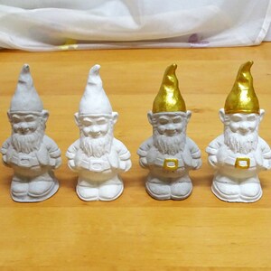 May include: Four white ceramic garden gnomes with gold hats. The gnomes are standing in a row on a wooden surface.