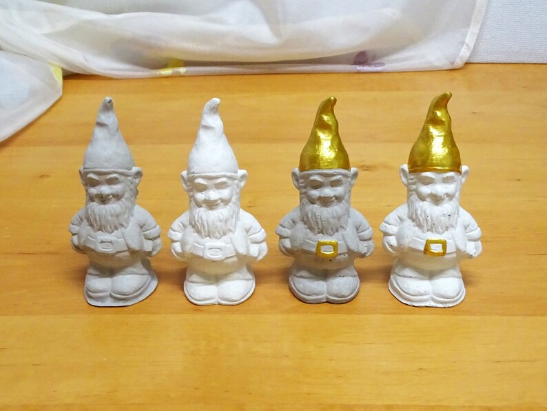 May include: Four white ceramic garden gnomes with gold hats. The gnomes are standing in a row on a wooden surface.