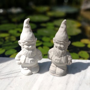 May include: Two white painted garden gnomes with long white beards and pointed hats. The gnomes are standing on a white surface.