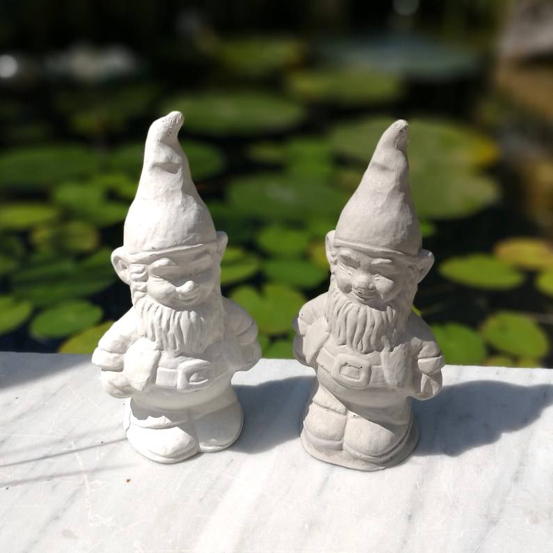 May include: Two white painted garden gnomes with long white beards and pointed hats. The gnomes are standing on a white surface.