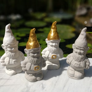 May include: Four white garden gnomes with gold accents. The gnomes are standing on a white surface.
