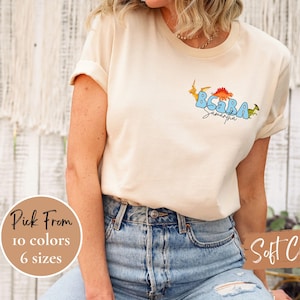Puede incluir: Camiseta color crema con un diseño de dinosaurio. El diseño incluye el texto "BCARA Samantha" en azul e ilustraciones de dinosaurios. La camiseta está disponible en 10 colores y 6 tallas. También se ve el texto "Soft Cream".