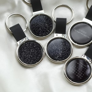 Könnte beinhalten: Eine Sammlung personalisierter Schlüsselanhänger mit einem schwarz-weißen Sternkarten-Design. Jeder Schlüsselanhänger hat einen runden, silberfarbenen Metallanhänger mit einem schwarzen Lederriemen und einem silbernen Schlüsselring. Einige Schlüsselanhänger sind mit Daten graviert.