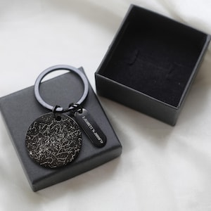 May include: A black keyring with a circular pendant displaying a white star map design. The keyring includes a rectangular tag with coordinates and is presented in a black gift box.
