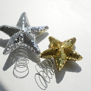 May include: Two star-shaped Christmas tree toppers. One is silver, the other is gold. Both are covered in small, reflective mirror tiles. Each star is attached to a silver metal spring base.
