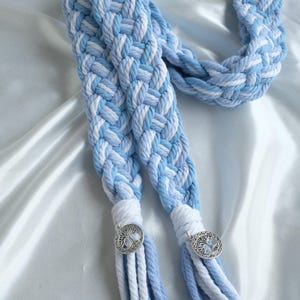 May include: A braided rope with blue and white stripes, tied with tassels and silver charms. The rope is on a light blue satin background.