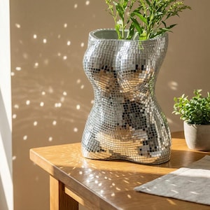 May include: A silver mosaic vase shaped like a torso, containing green plants. The vase is constructed from small, reflective tiles, giving a disco ball appearance. It rests on a wooden table, with a small potted plant and a linen napkin close by.