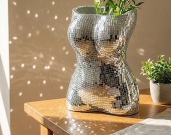 May include: A silver mosaic vase in the shape of a torso, holding green plants. The vase is made of small, reflective tiles, creating a disco ball effect. It sits on a wooden table, with a small potted plant and a linen napkin nearby.